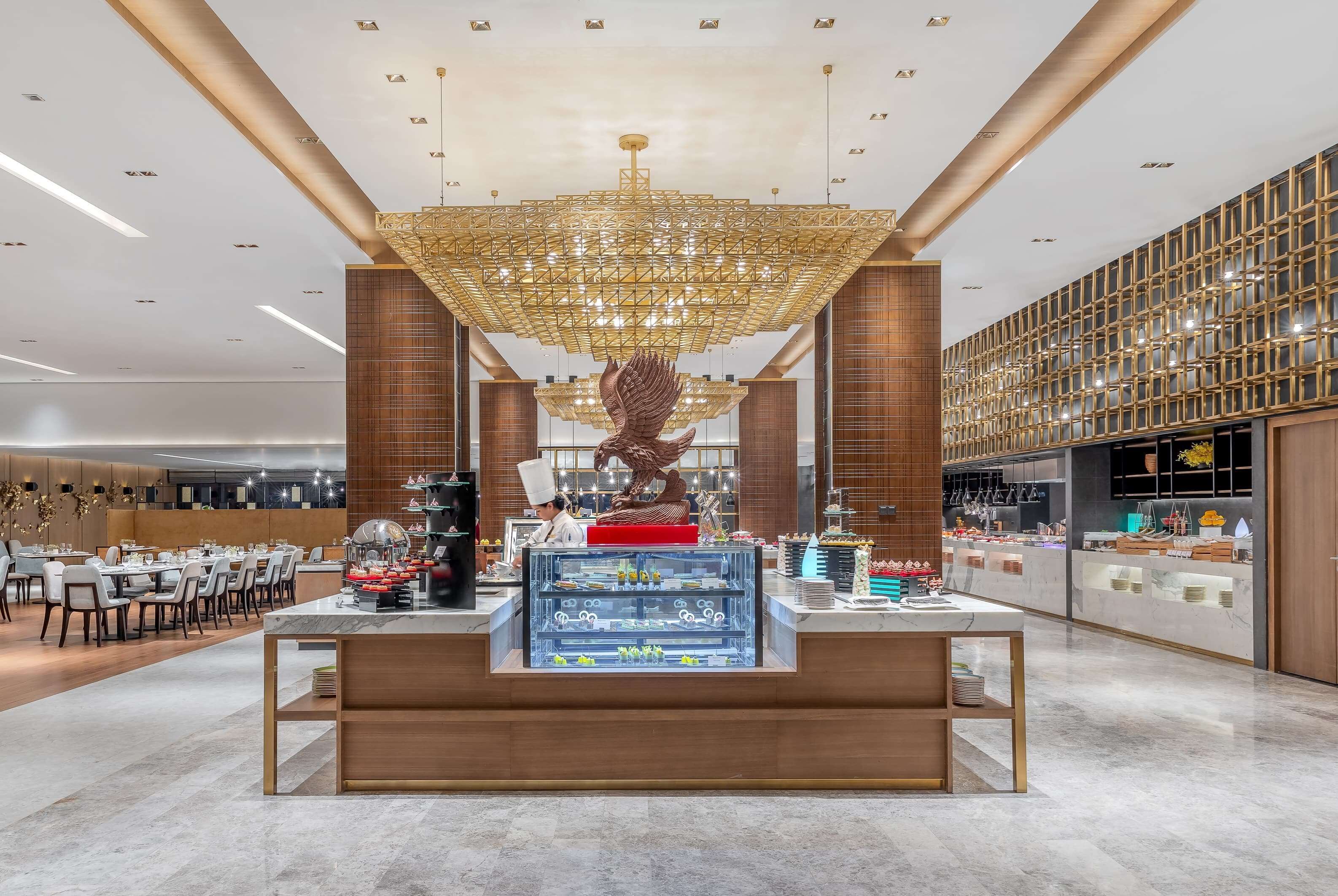 A modern interior with a central dessert display, a large chandelier, and a patterned wall.