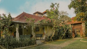 A house with a tiled roof, a porch, and a garden with various plants and a bench. - Villa Chempaka (Ubud)