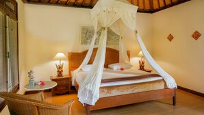 A bedroom with a four-poster bed, wooden headboard, and a ceiling fan. There is a nightstand with a lamp and a small statue. A wicker chair is placed near the bed. - Villa Chempaka (Ubud)
