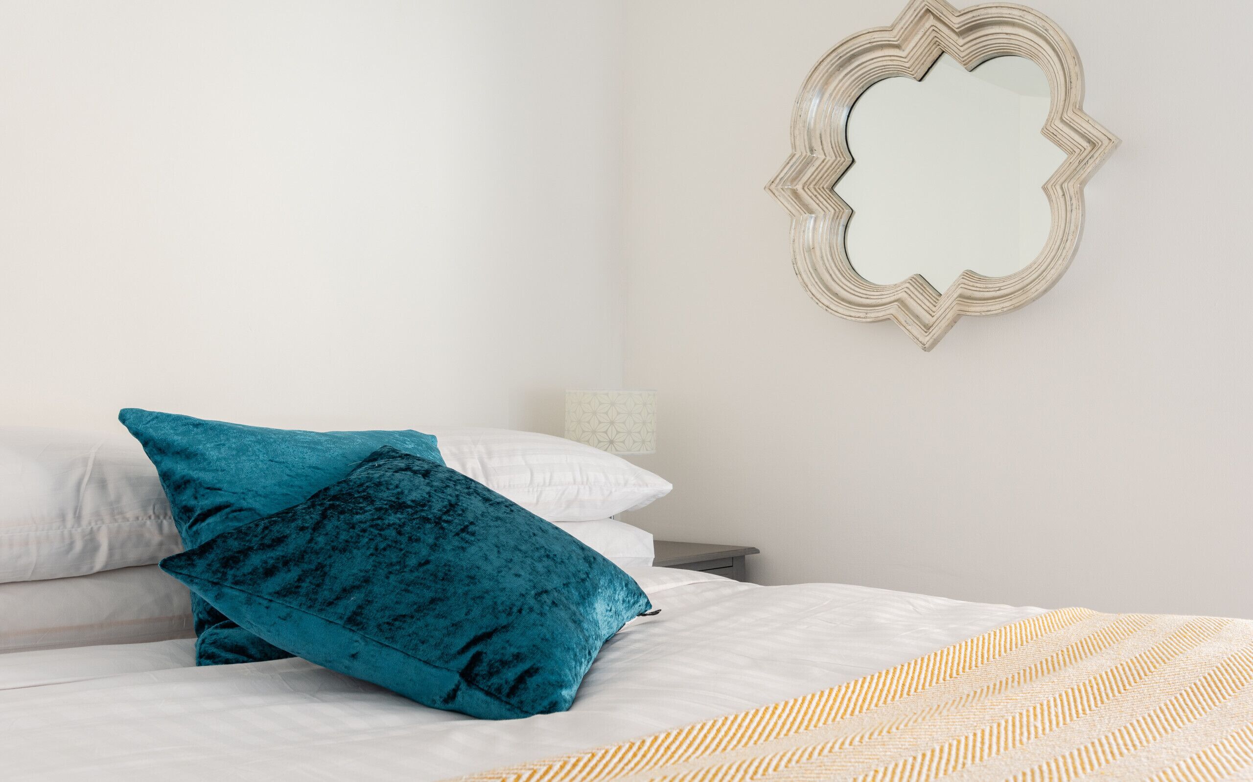 Una cama bien hecha con sábanas blancas y dos almohadas color turquesa, un espejo decorativo en la pared y una manta estampada.
