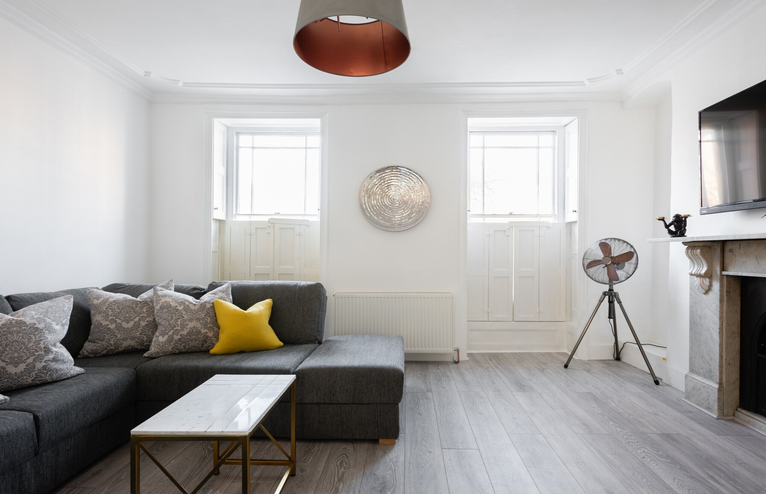 Una sala moderna con un sofá seccional gris, una mesita y un ventilador. Hay dos ventanas con persianas blancas y una chimenea sobre la cual se encuentra un televisor.
