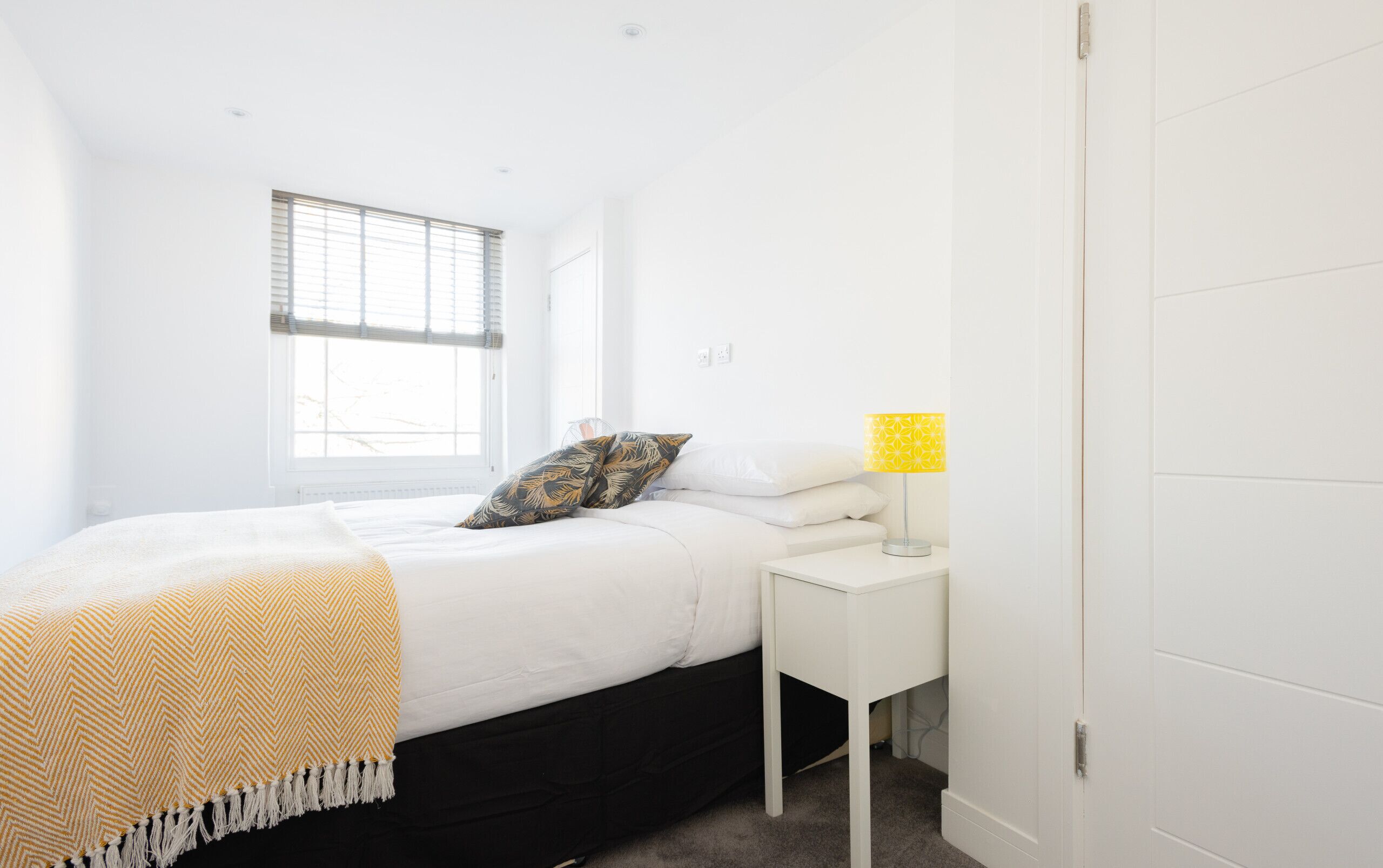 A bedroom with a bed, a bedside table with a lamp, a window with blinds, and a door.