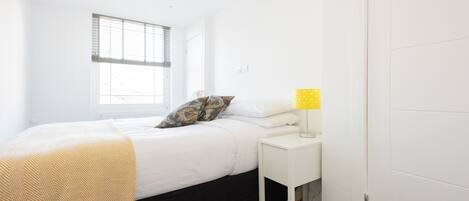 A bedroom with a bed, a bedside table with a lamp, a window with blinds, and a door.
