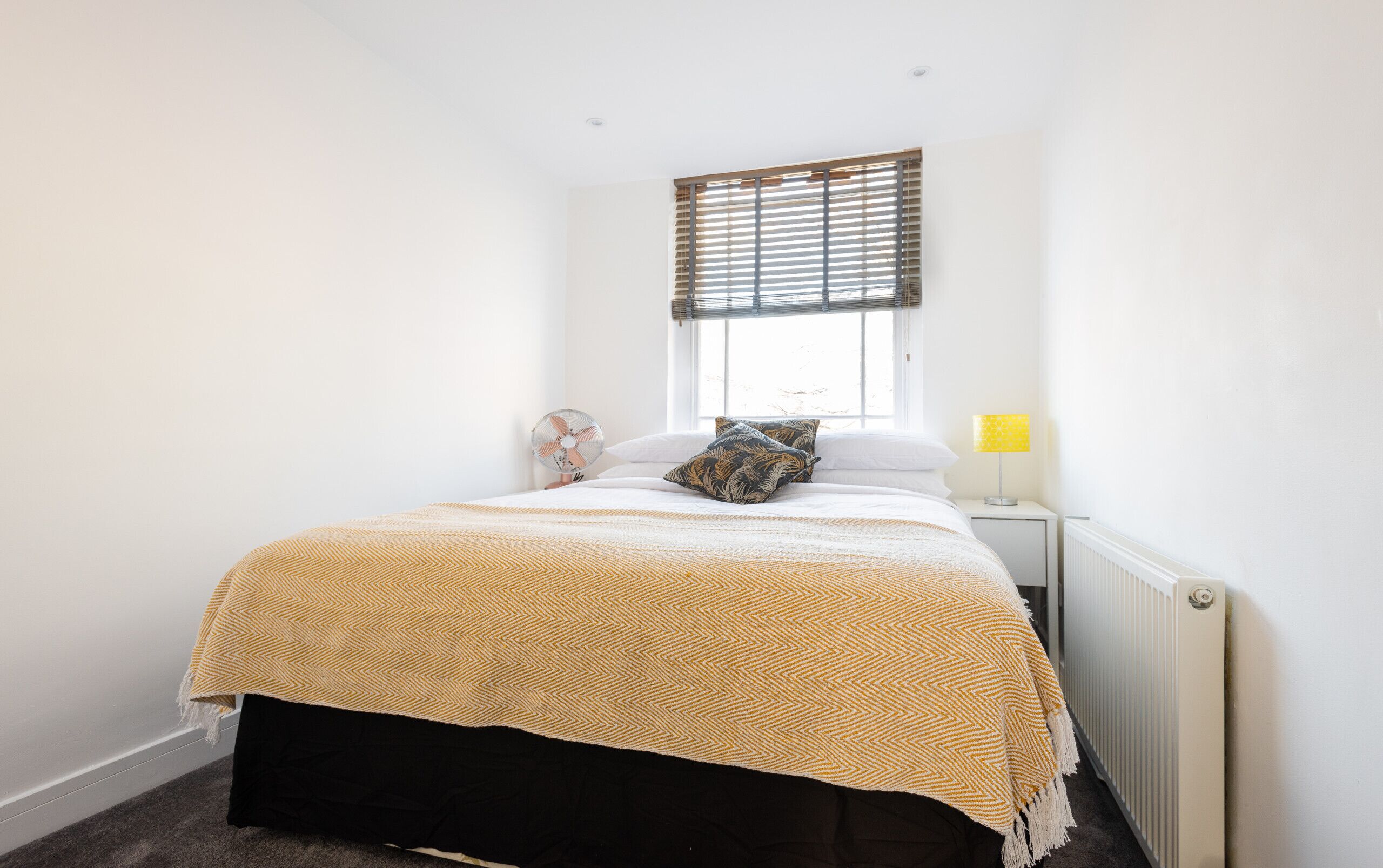 Quarto com cama, mesa de cabeceira com luminária, janela com persianas e radiador.