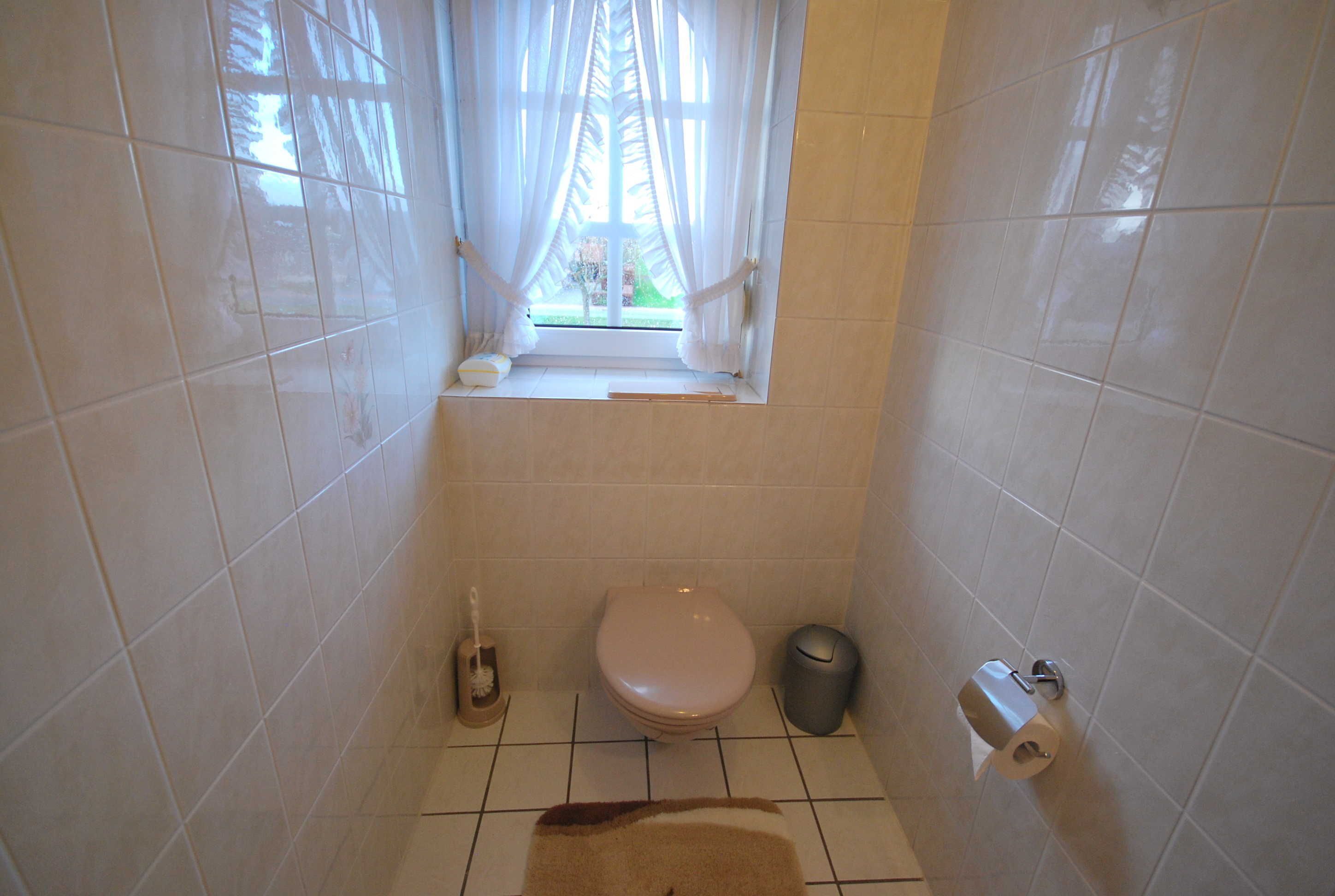 A small bathroom with a toilet, a trash bin, a toilet paper holder, and a window with curtains.