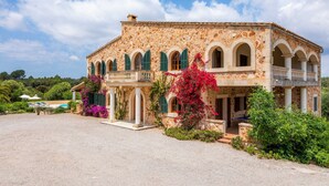 A stone building with arched windows and a balcony, surrounded by greenery and a clear sky.
