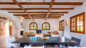 A dining area with a large table, chairs, and a fruit bowl. There are arched windows and a fireplace.