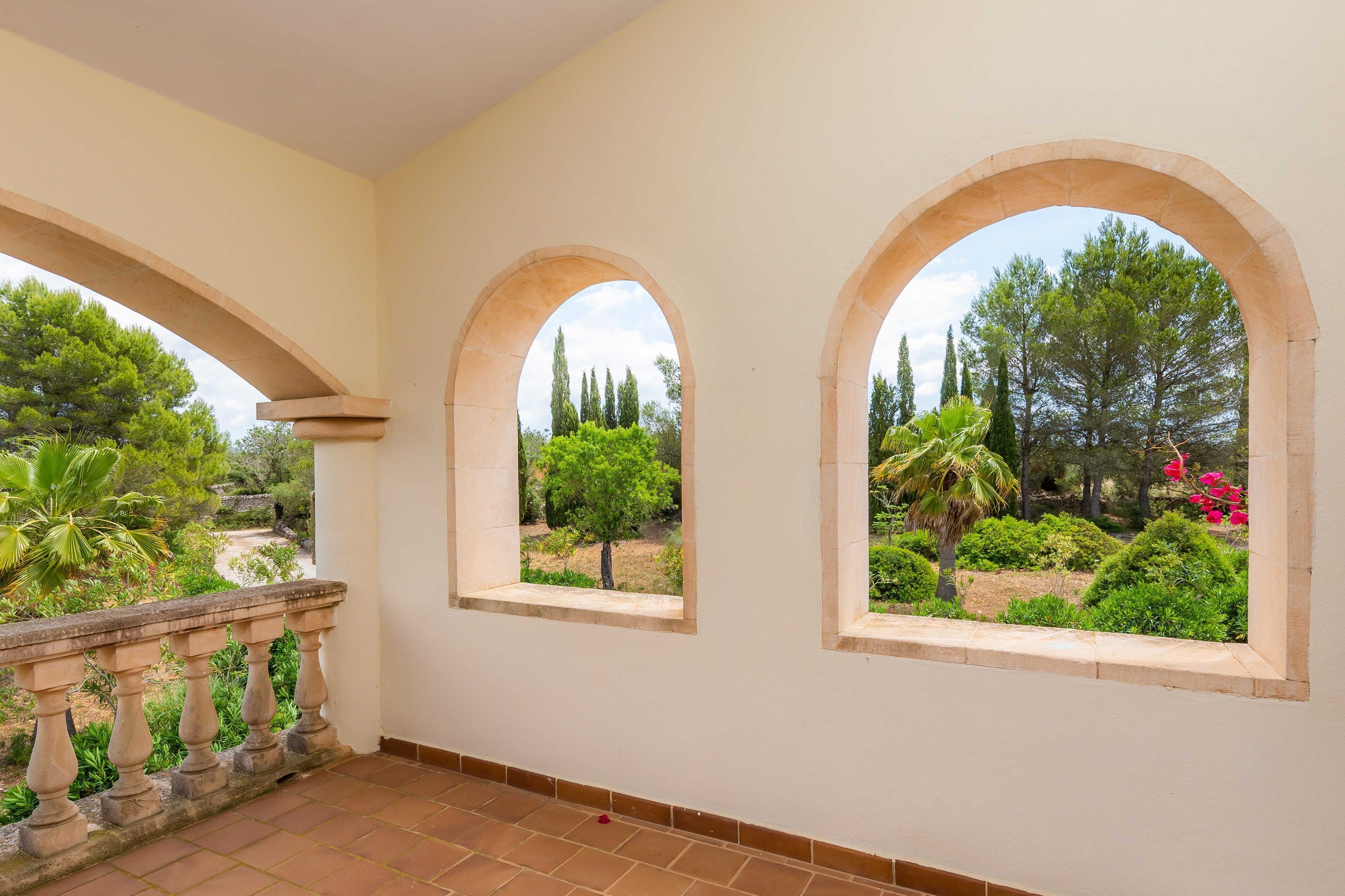 A balcony with a tiled floor, a stone balustrade, and two arched windows overlooking a garden with trees and shrubs.