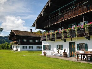 A traditional chalet with a balcony adorned with flowers, a wooden deck, and a view of a lush green field. - Ferienwohnung "In der Stube"  (28qm) (Kiefersfelden)