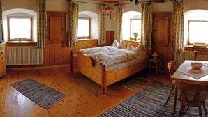 A cozy bedroom with a wooden bed, a small table with a chair, and a window with curtains. - Ferienwohnung "In der Stube"  (28qm) (Kiefersfelden)