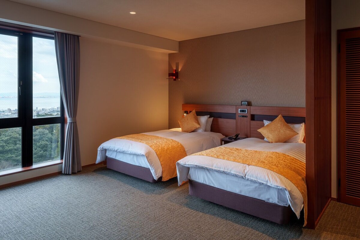 A hotel room with two beds, a large window with a view, a wall-mounted TV, and a wooden headboard.