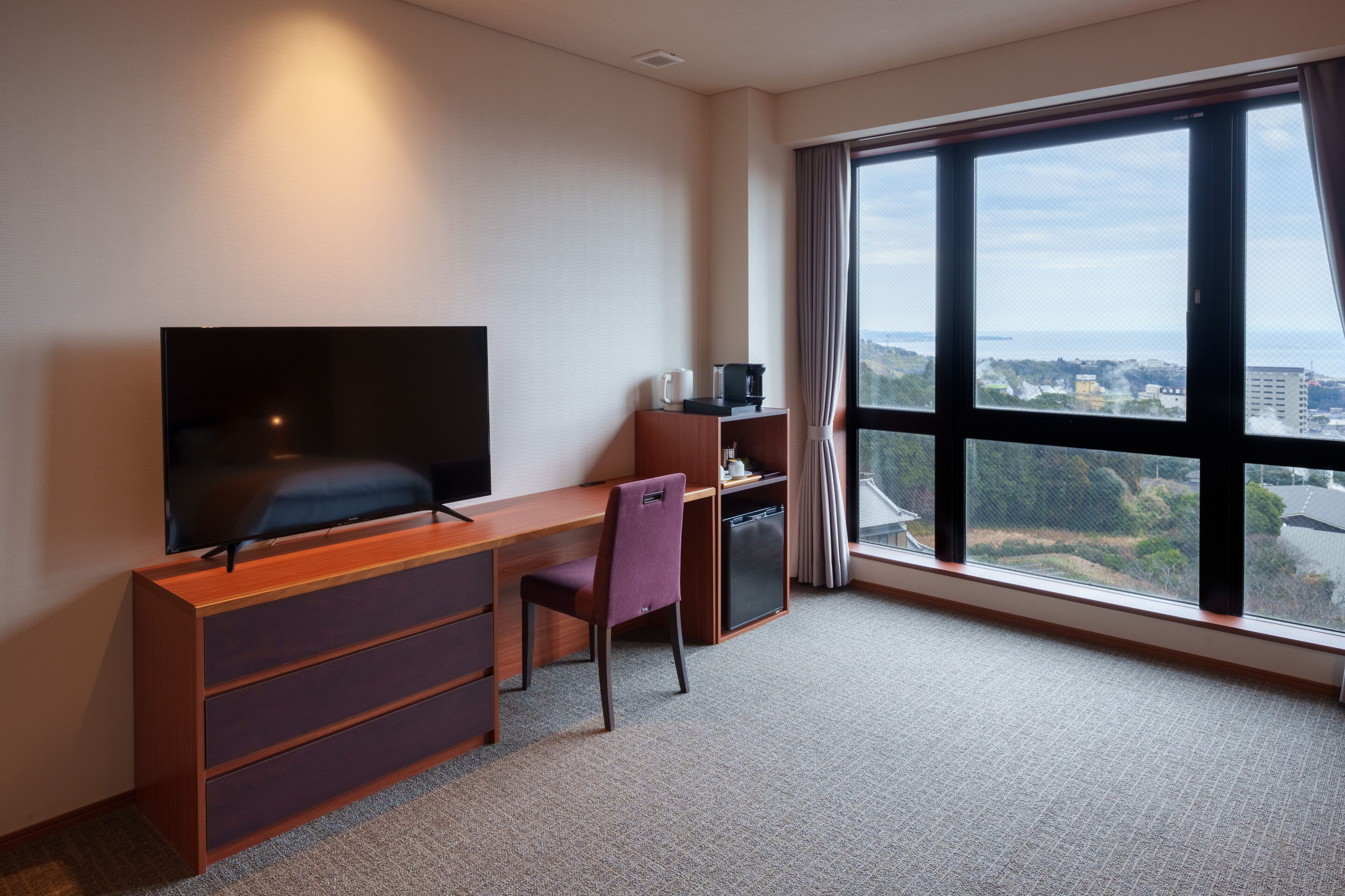 Habitación de hotel con un ventanal grande, un televisor de pantalla plana, un escritorio con silla y un mueble auxiliar pequeño.