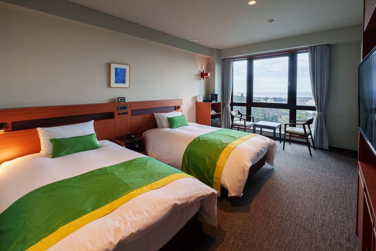 A hotel room with two beds, a desk, a chair, a TV, and a large window with a city view.