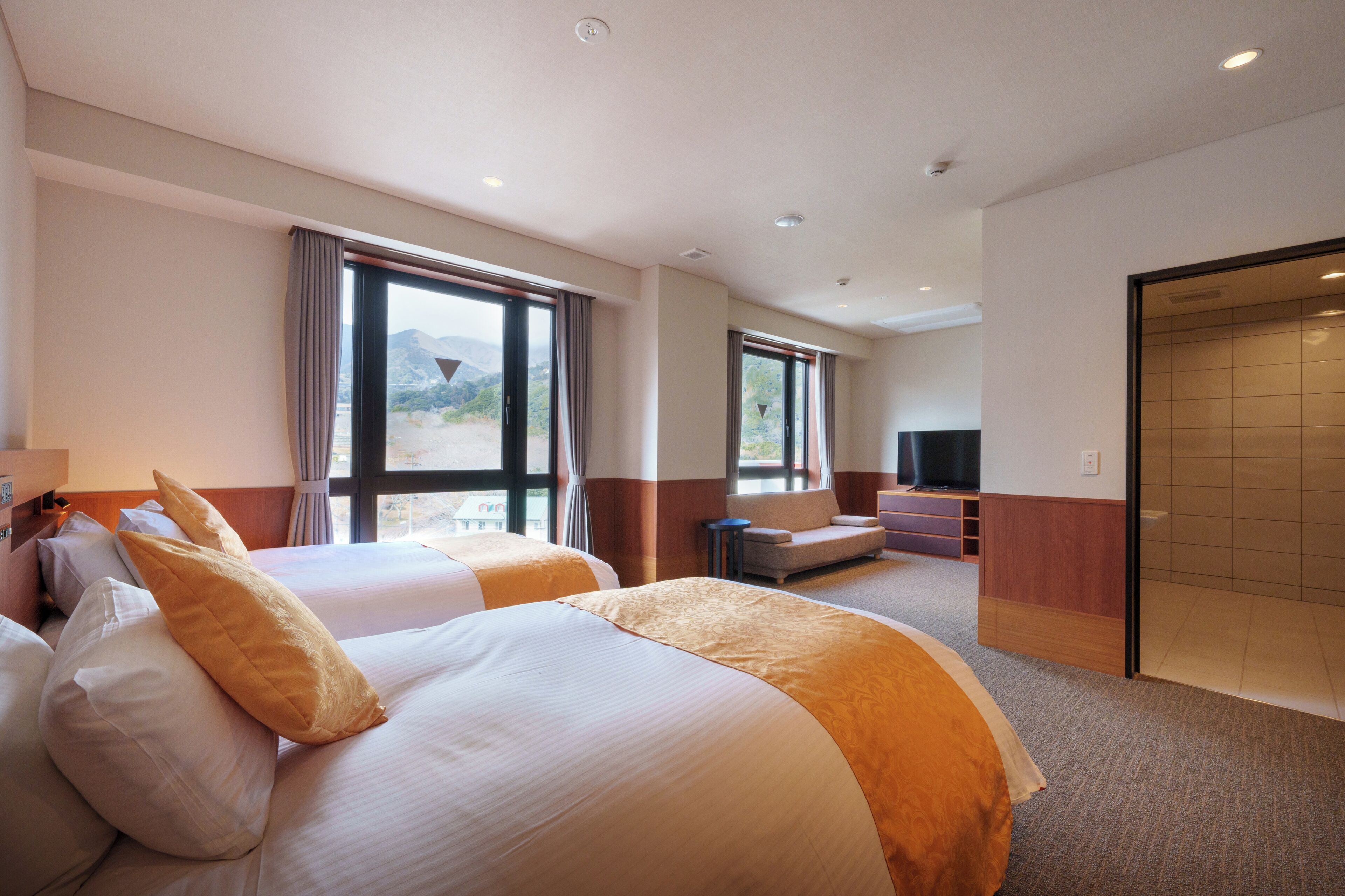 A hotel room with two beds, a sofa, a TV, and large windows offering a view of the outdoors.