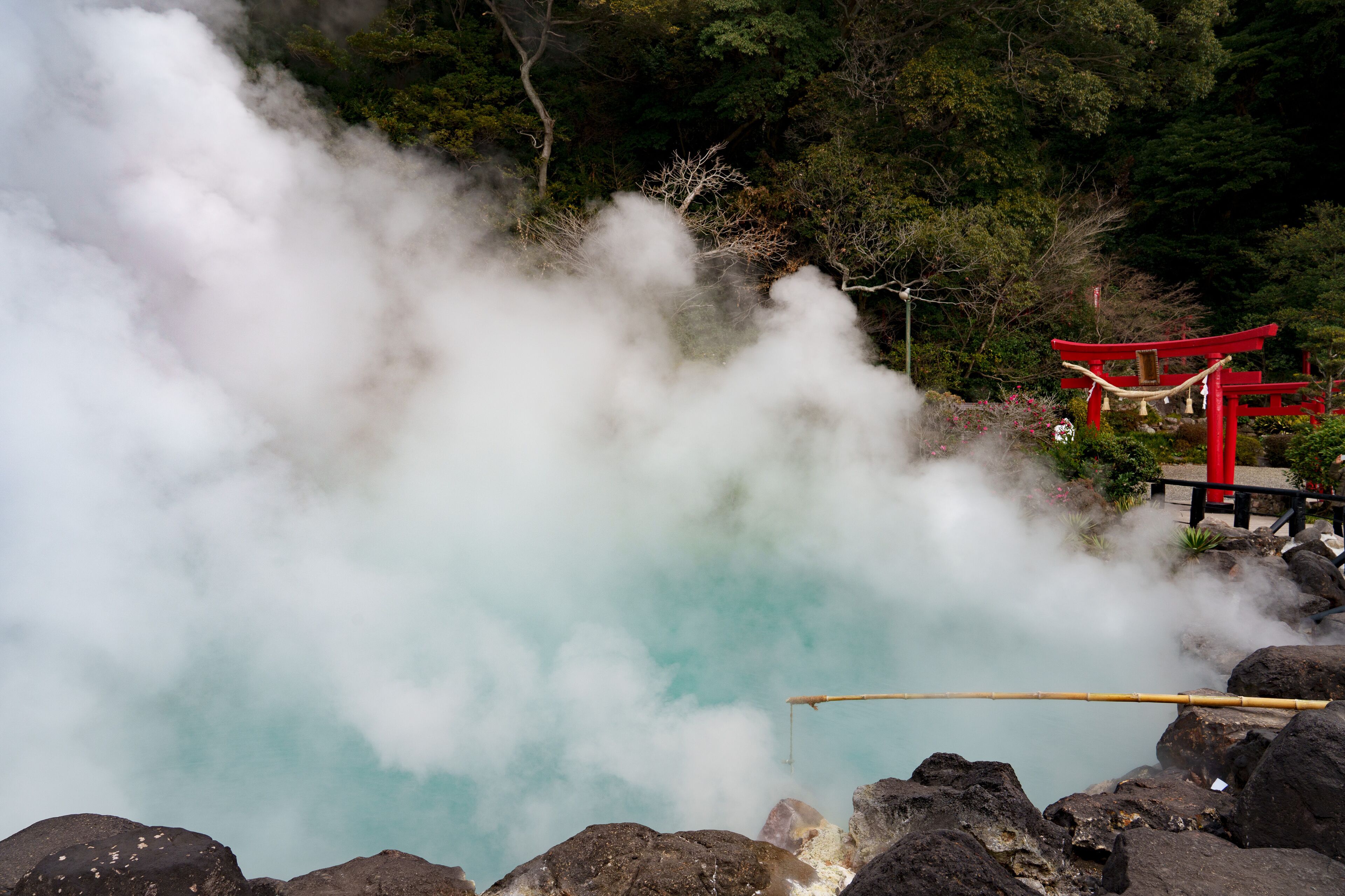 A hot spring with steam rising from the water, surrounded by rocks and a red wooden structure.