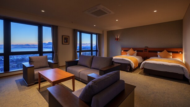 A hotel room with a large window offering a city view, a sofa, a bed, and a small table.