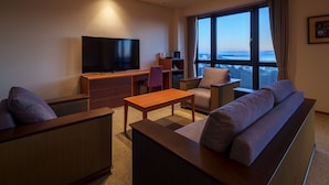 A modern hotel room with a large flat-screen TV, a sofa, a wooden coffee table, and a view of the city through large windows.