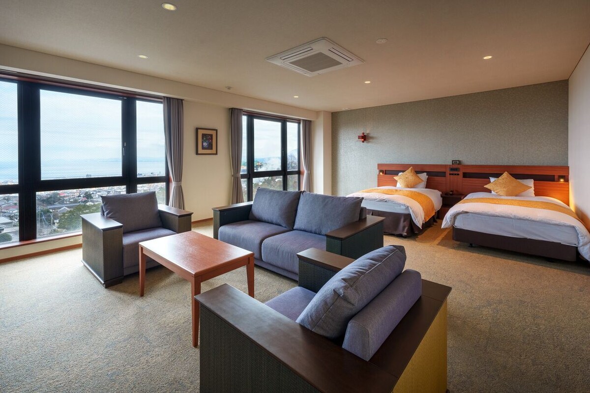 A hotel room with a sofa, two single beds, a small table, and large windows offering a city view.