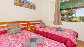 Quarto com duas camas, cabeceira de madeira, mesa de cabeceira, janela com vista e um quadro na parede.