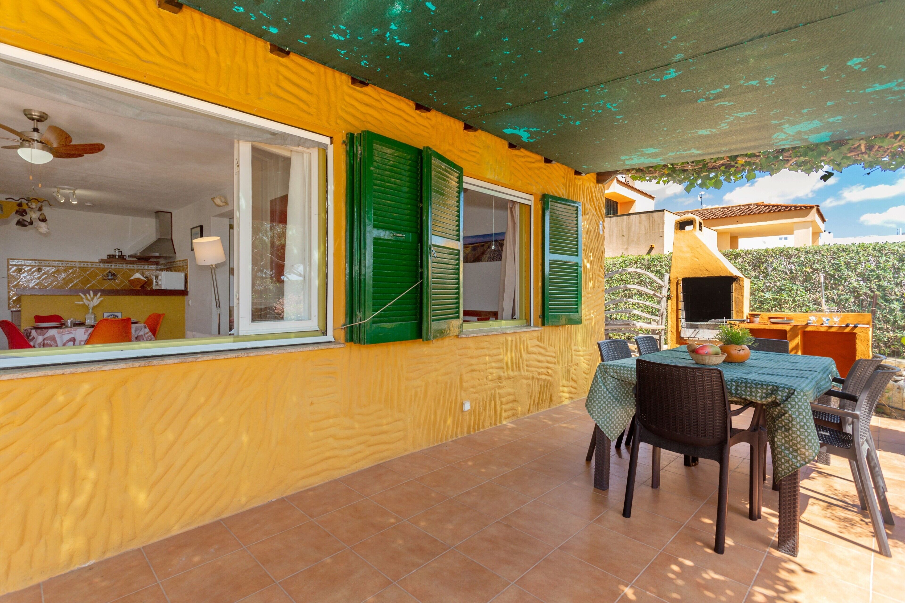 A yellow exterior wall with green shutters, a tiled patio with a table and chairs, and a visible kitchen area.