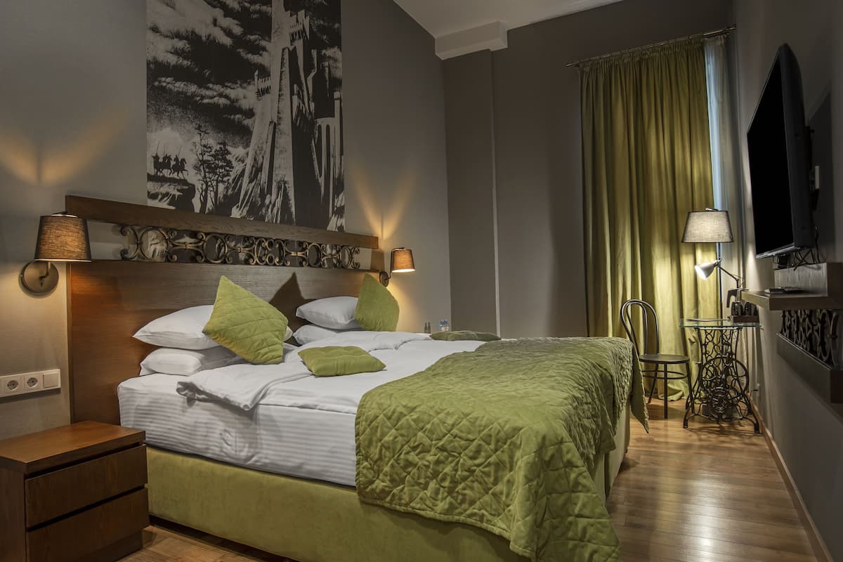 Een netjes opgemaakt bed met een groene sprei, twee bedlampen, een houten nachtkastje, een stoel, een tafel met een lamp, en een groot schilderij aan de muur.