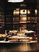 Zona de bar con estantes llenos de botellas, mostrador con pasteles y una lámpara decorativa.