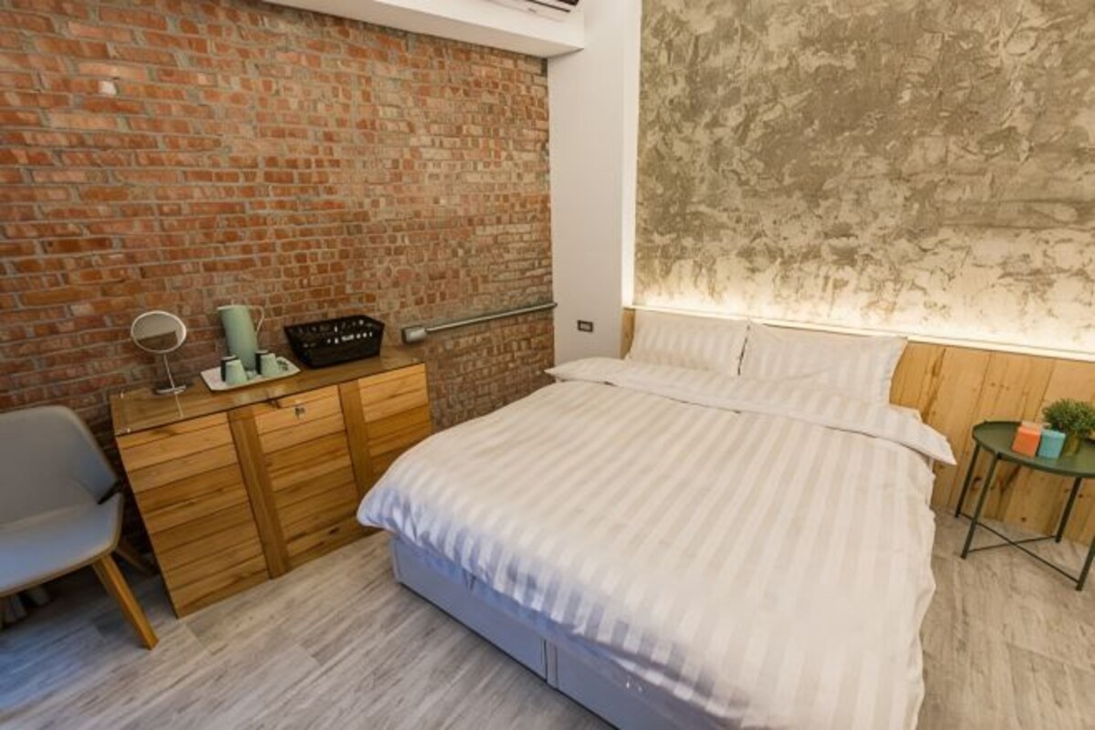 Una camera da letto con parete in mattoni, comò in legno, letto e un piccolo tavolo con una lampada.