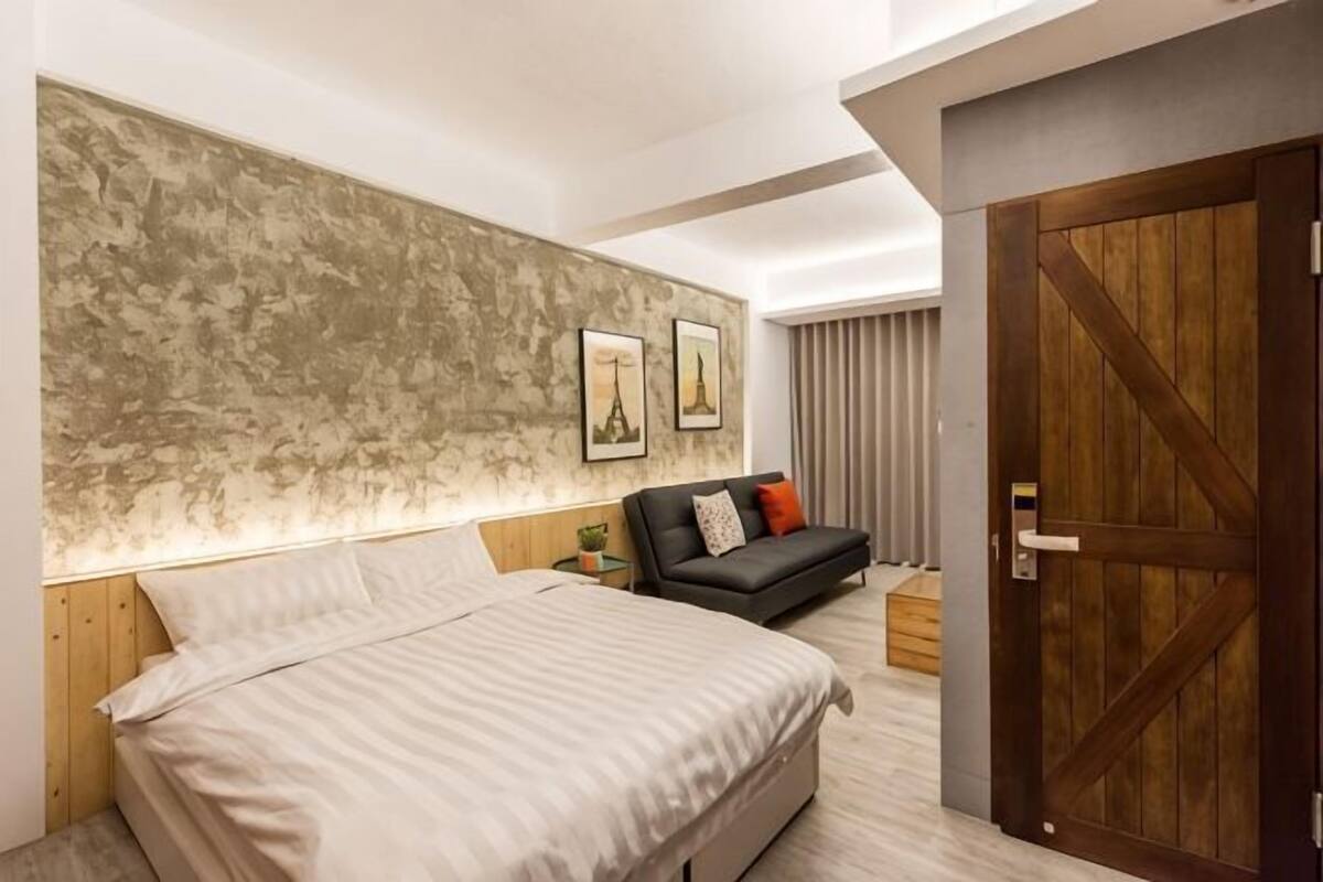 A bedroom with a bed, a sofa, a wooden door, and a textured wall.