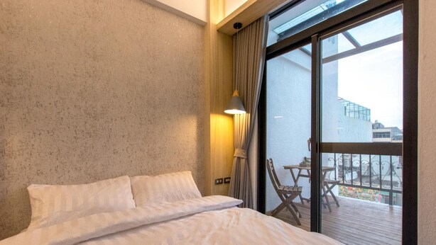 Una habitación de hotel con una cama grande, un ventanal con cortinas y vistas a un balcón con mobiliario exterior.