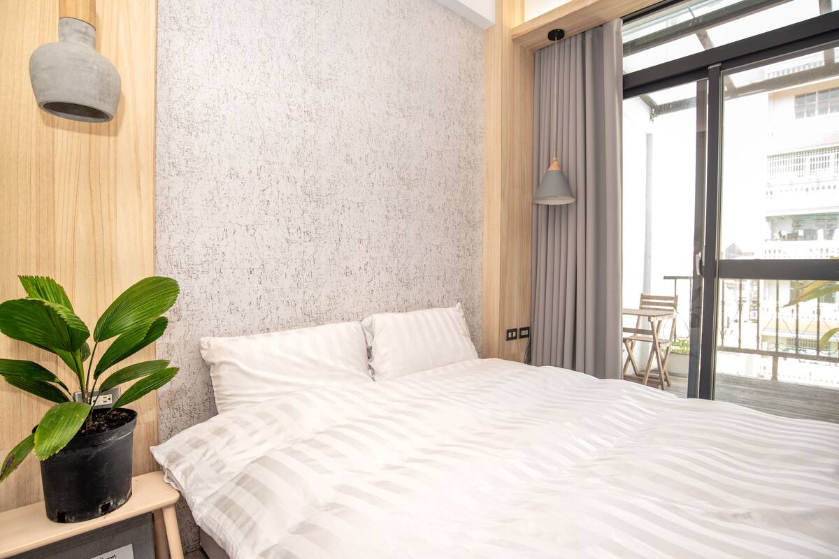 A modern hotel room with a large bed, a potted plant, a wooden nightstand, and a view of the city through a large window.