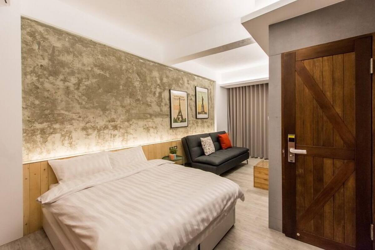 A modern bedroom with a large bed, a dark sofa, and a wooden door.