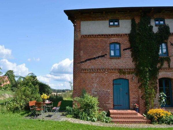 A brick building with a blue door, a small porch with a table and chairs, and a garden with flowers and shrubs.