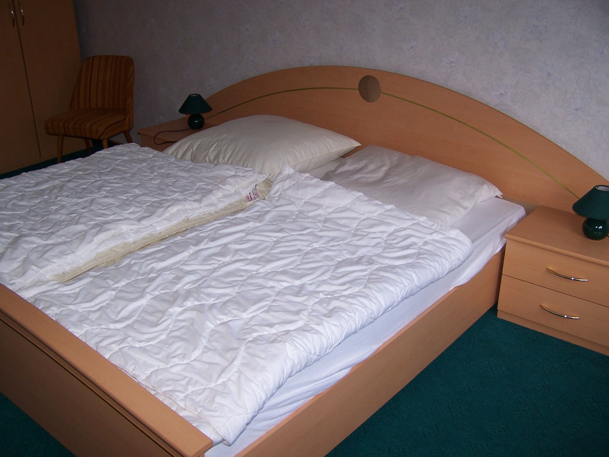 Una cama con ropa de cama blanca, un cabecero de madera, una mesita de noche con lámpara y una silla.