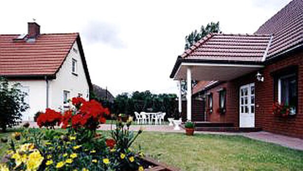 Una zona residencial con dos casas, una de techo rojo y otra con fachada blanca. Hay un jardín con flores amarillas y rojas frente a las casas.