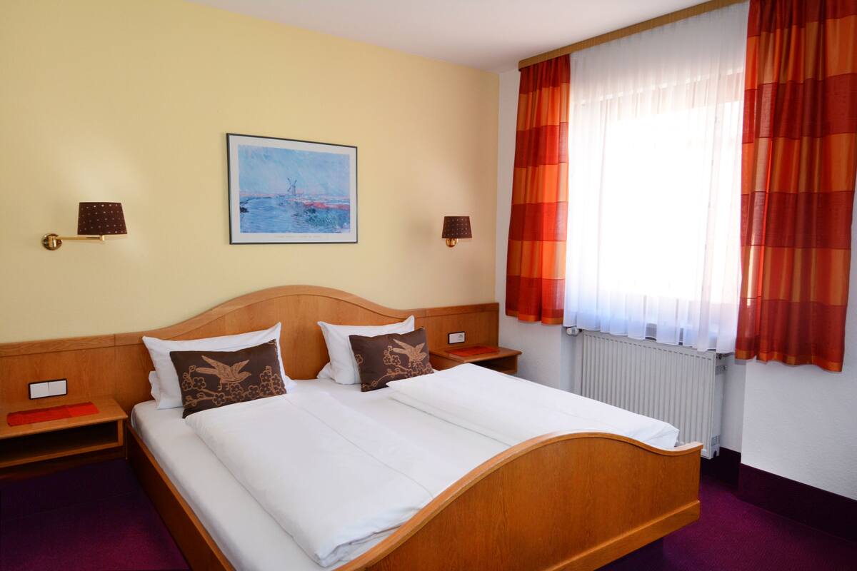 A hotel room with a wooden bed, two bedside tables, a nightstand, a framed picture on the wall, and a window with curtains.