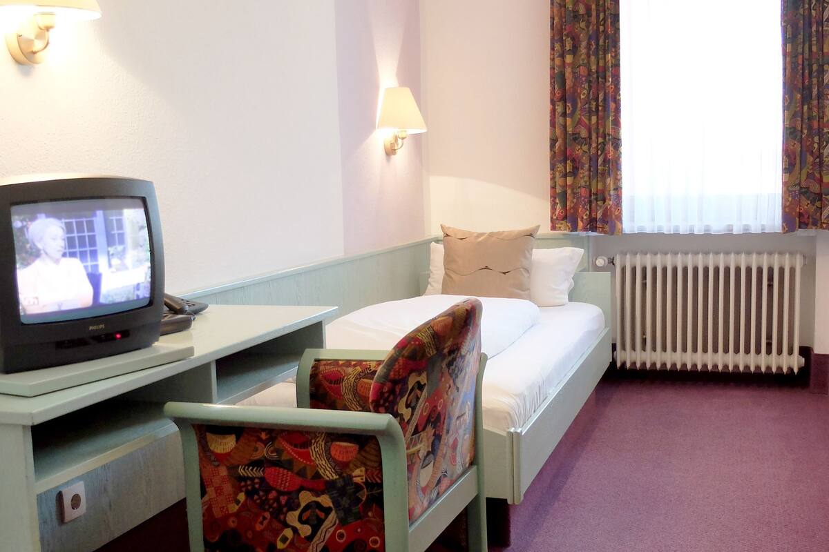 A hotel room with a bed, a desk with a television, a chair, a window with curtains, and a radiator.