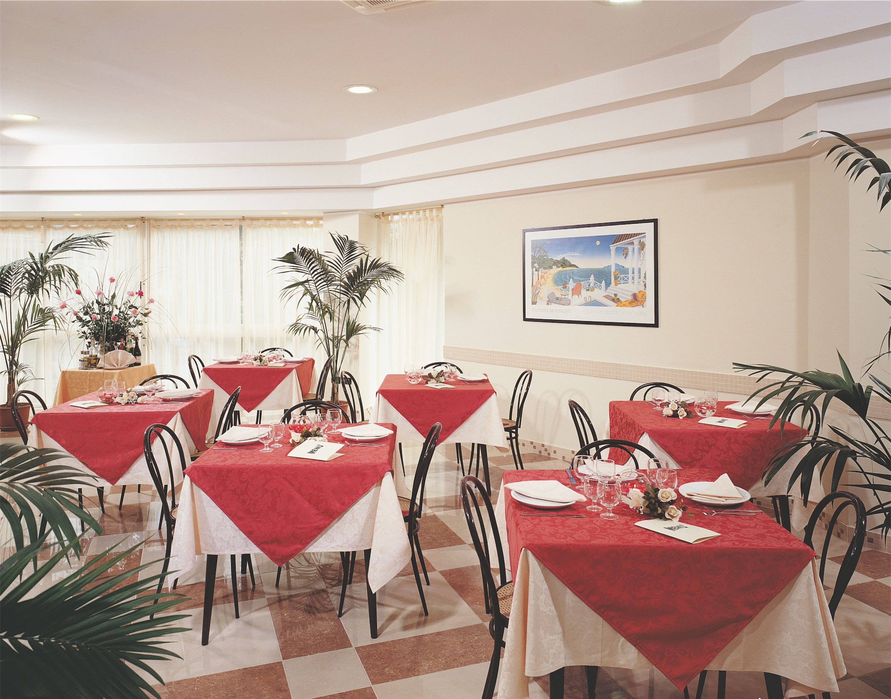 A dining area with tables set for four, each with a red tablecloth, white plates, and glasses. There are potted plants and a painting on the wall.