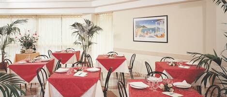 A dining area with tables set for four, each with a red tablecloth, white plates, and glasses. There are potted plants and a painting on the wall.