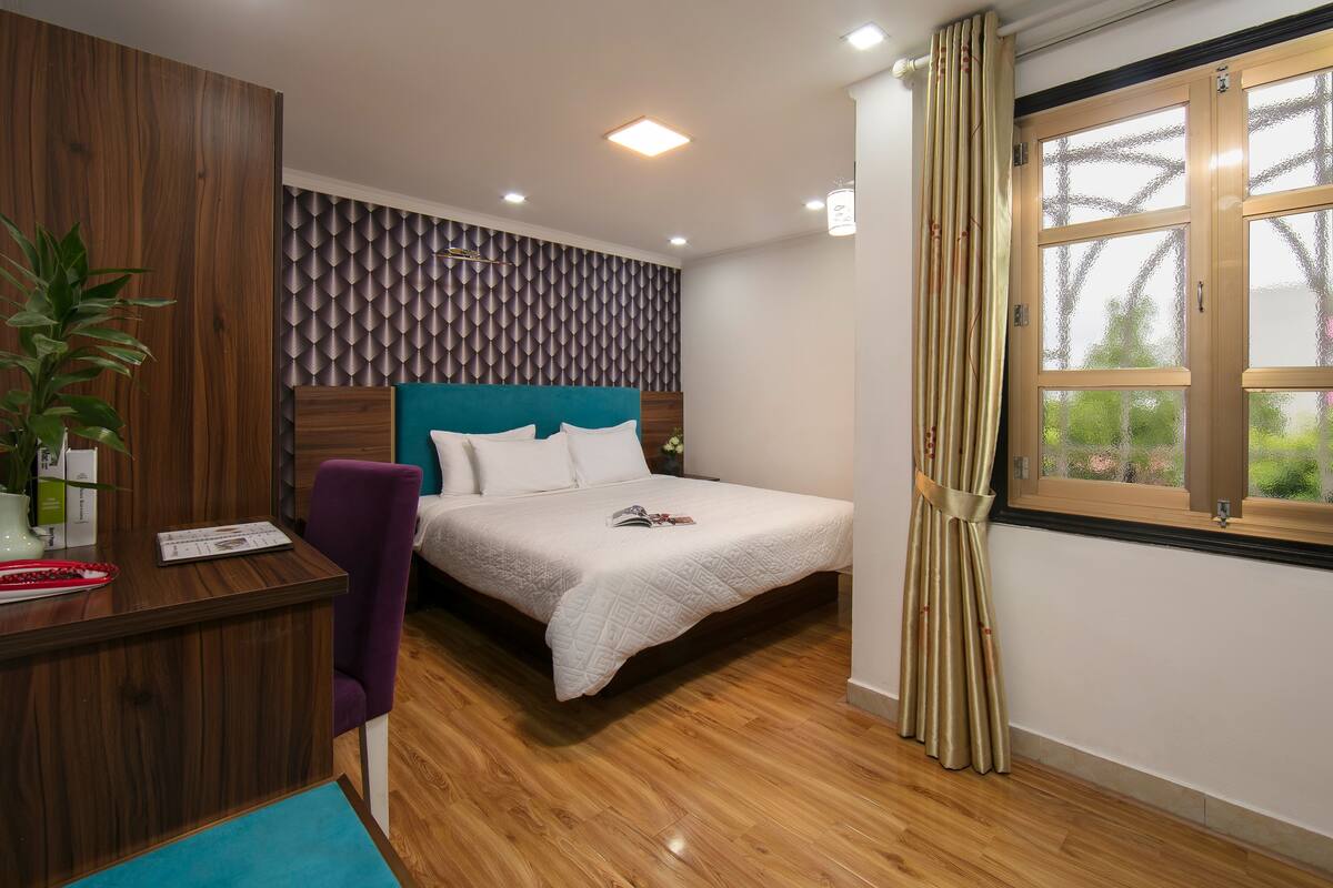 A bedroom with a wooden desk, a purple chair, a bed with a white quilt, and a window with curtains.