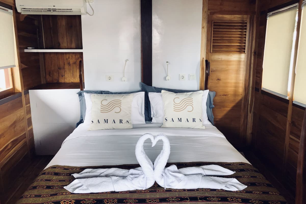 Una habitación tipo cabaña de madera con una cama, dos almohadas bordadas con 'SAMARA' y una toalla blanca doblada en forma de cisne, además de una ventana con persianas.