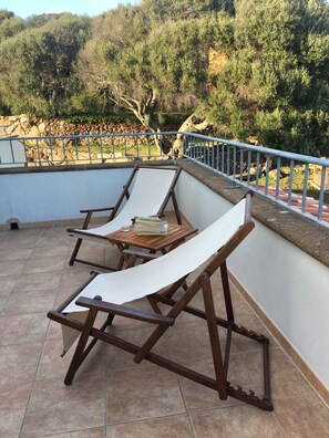 Una terraza con dos sillones de madera y una mesita, rodeada por un muro de piedra y una barandilla de metal.
