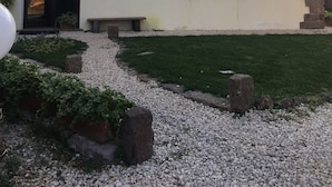 A two-story building with a white facade, stone accents, a black door, and a small garden area with gravel and plants. - Artemisia Sardegna (Tergu)