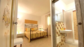 A bedroom with a bed, bedside table, and chair. There is a bathroom with a sink and mirror. - Artemisia Sardegna (Tergu)