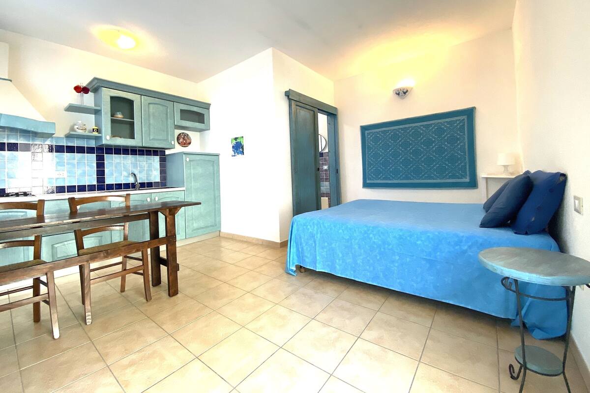 Quarto pequeno com cama, uma mesinha, cozinha compacta e porta do banheiro.