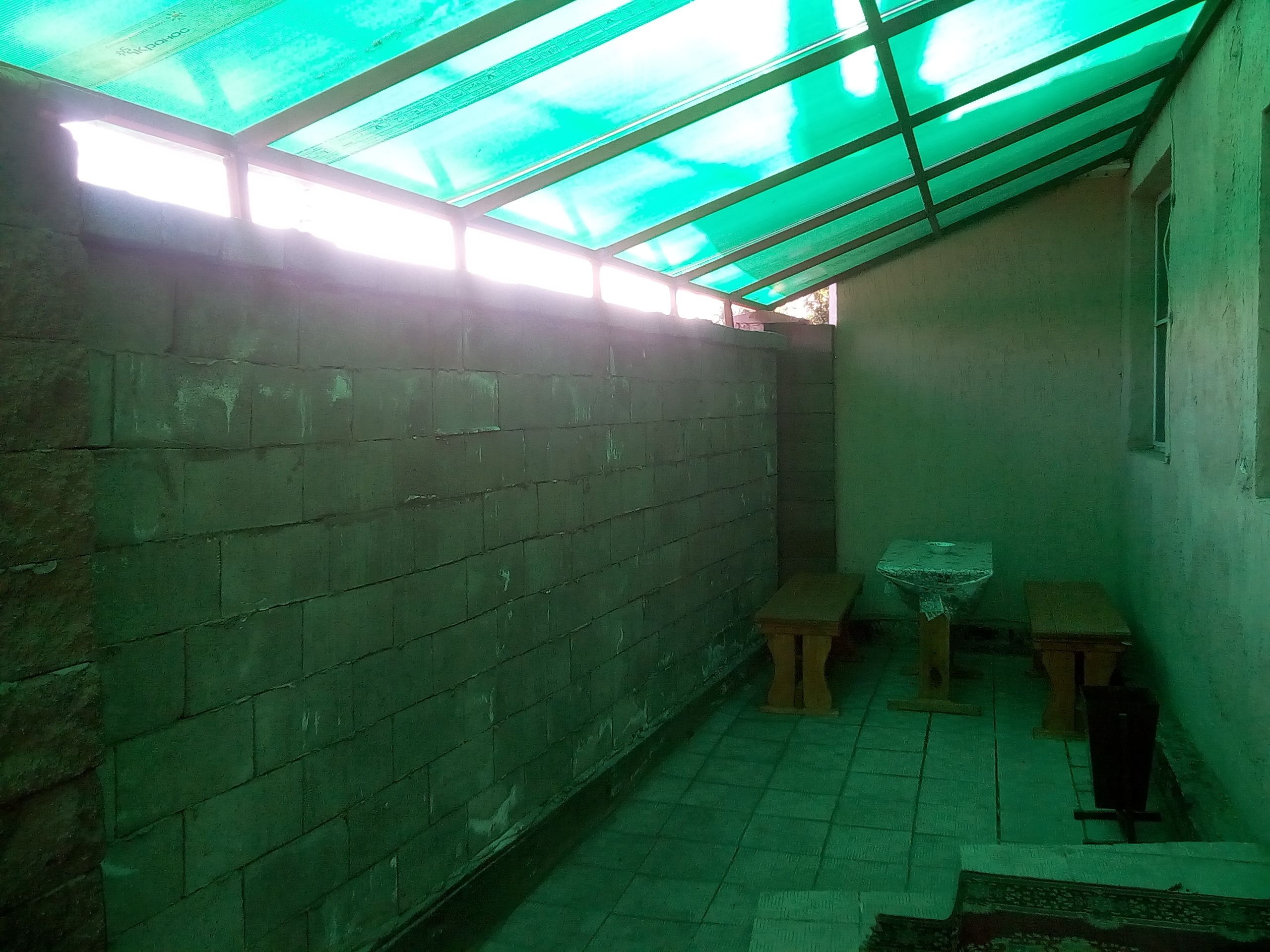 Ein kleiner, abgeschlossener Raum mit Fliesenboden, Ziegelwand und einem grün schimmernden Dach. In der Ecke steht ein kleiner Tisch und ein Hocker.