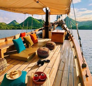 A wooden deck with seating, a table with drinks, and a view of green hills. - Samara II Liveaboard (Labuan Bajo)