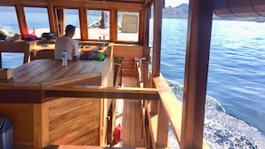 Un bateau en bois avec une table et des chaises, une personne assise à la table, et une vue sur l’eau.