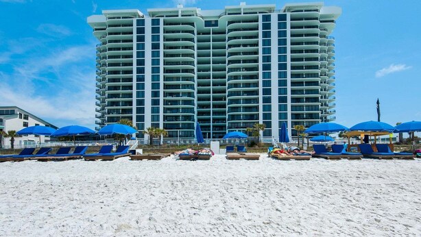 Ein Hochhaus-Hotel mit mehreren Balkonen, ein Sandstrand im Vordergrund und blaue Sonnenschirme.