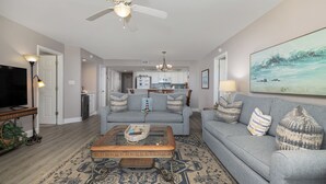 A living room with a sofa, coffee table, and TV. There is a kitchen in the background. - Stay @ Sandy Pause on Folly Beach (Folly Beach)