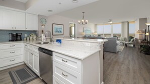 A spacious kitchen with white cabinets, a dishwasher, and a large island. There is a dining area with a sofa and chairs, and a view of the ocean through large windows. - Stay @ Sandy Pause on Folly Beach (Folly Beach)